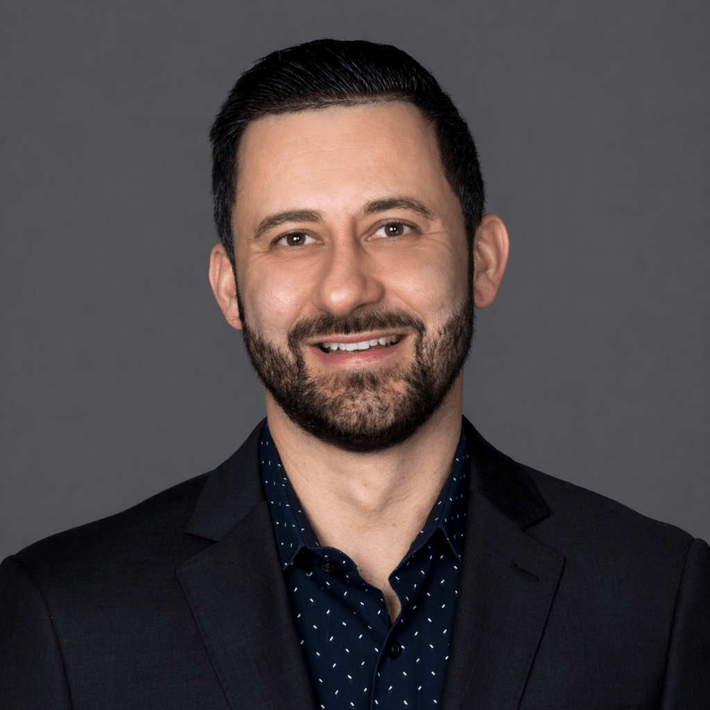 Head-and-shoulders portrait of a smiling man with dark hair and a beard, wearing a black blazer and navy shirt, against a gray studio backdrop.
