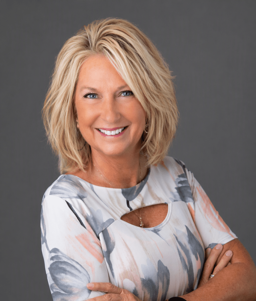 Smiling woman with short blonde hair in a floral blouse stands with arms crossed against a gray studio backdrop.