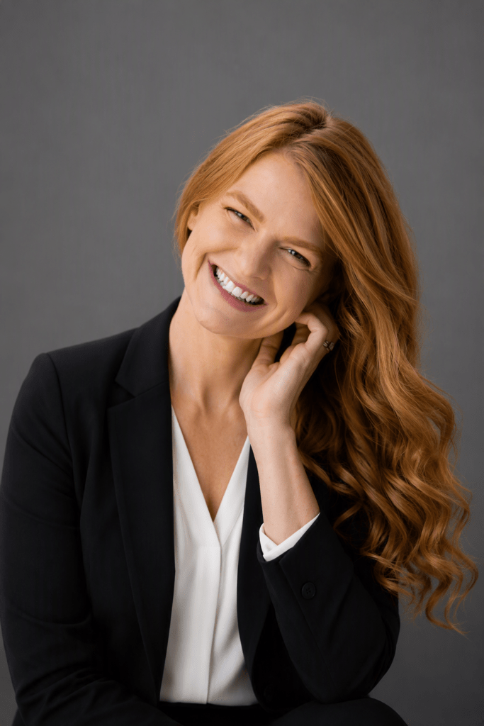 Professional headshot of a smiling woman with long red hair in a black blazer and white blouse against a gray backdrop.