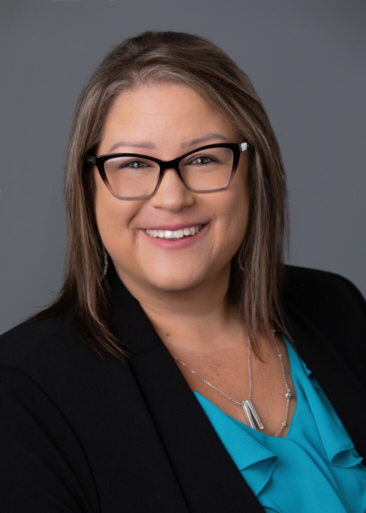 Portrait of a smiling woman with glasses, wearing a black blazer and turquoise blouse.