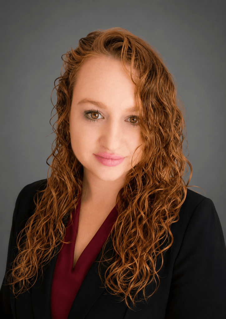 Professional headshot of a woman with curly red hair, wearing a black blazer and burgundy blouse, against a gray backdrop.