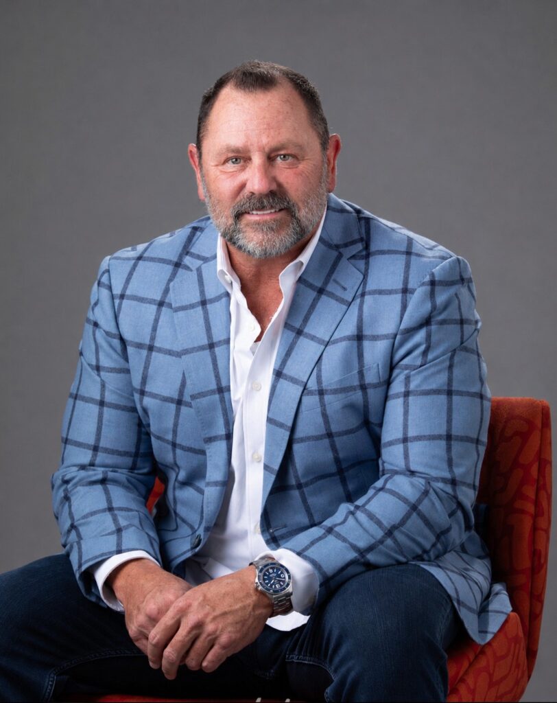 Man in a light blue plaid blazer and white shirt, seated on an orange chair, smiling against a gray backdrop.