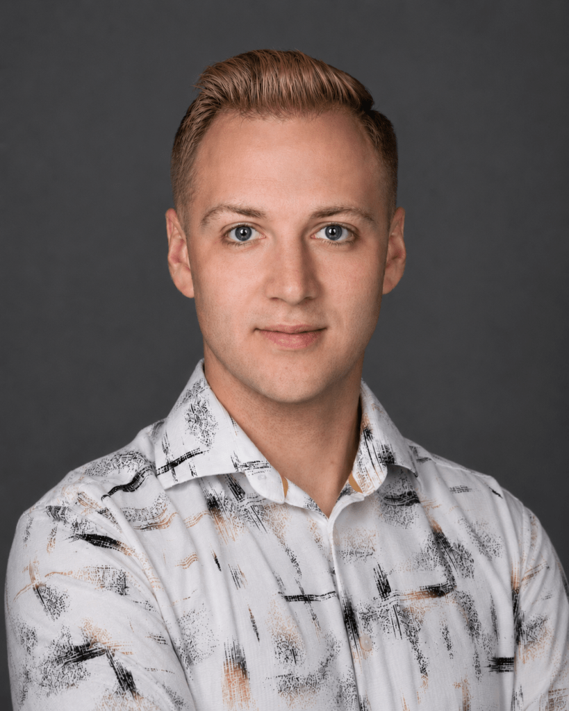 Head-and-shoulders portrait of a young man with blond hair and blue eyes, wearing a white patterned shirt against a gray studio background.