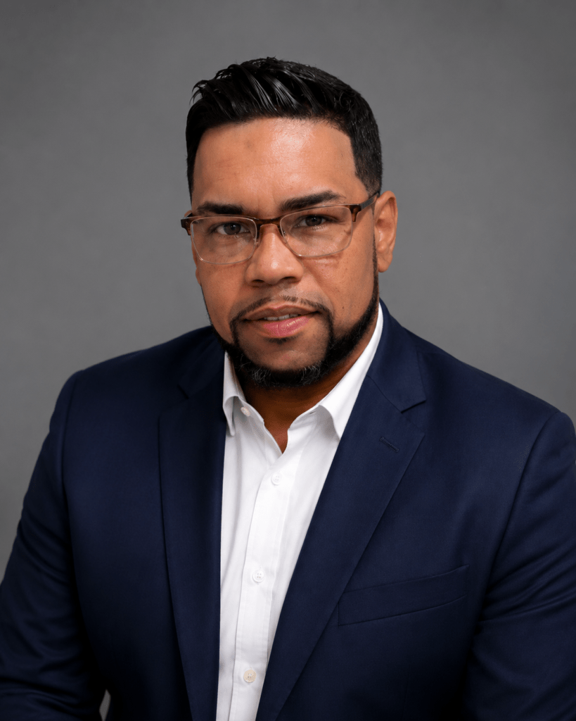Professional headshot of a man with dark hair and glasses, wearing a navy blazer and white shirt against a gray studio backdrop.