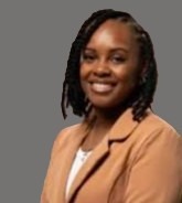 Smiling woman with short dreadlocks wearing a tan blazer and white top, against a gray background.
